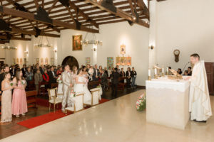 Padre en la iglesia dando el sermón a los futuros esposos mientras ellos lo miran de pie, acompañados de sus invitados.