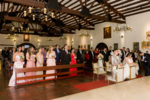 Pareja de novios en el altar, con su familia acompañándolos en la iglesia vestidos elegantes esperando la bendición del padre