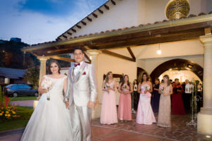 Mujer con vestido de novia, con su esposo al lado a la salida de la iglesia, detrás de ellos sus damas de honor mientras cae la tarde