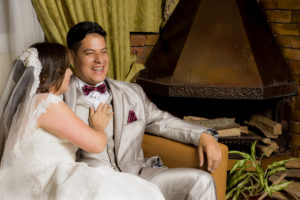 Pareja de esposos al lado de chimenea en la sala riéndose, ella con un vestido de novia blanco y el vestido con un traje color gris brillante.