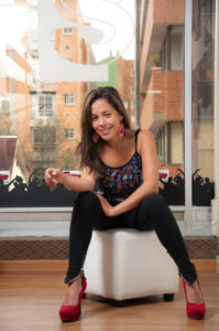 Mujer con cabello suelto y tacones, sentada en puff, sonriente.
