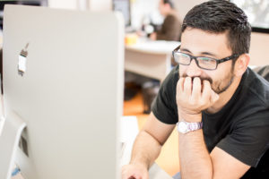 Hombre sonriente con gafas y barba con una mano en la boca recargado en la mesa mientras mira la pantalla del computador Mac