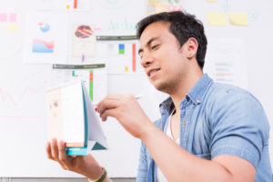 Hombre vestido con camisa de jeans y camisa blanca analizando un calendario en la oficina, con estadísticas en un tablero de fondo.