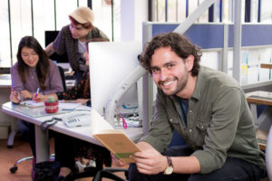 Hombre sentado en oficina de camisa verde mirando a la cámara con un libro en las manos, en el fondo estudiantes conversando