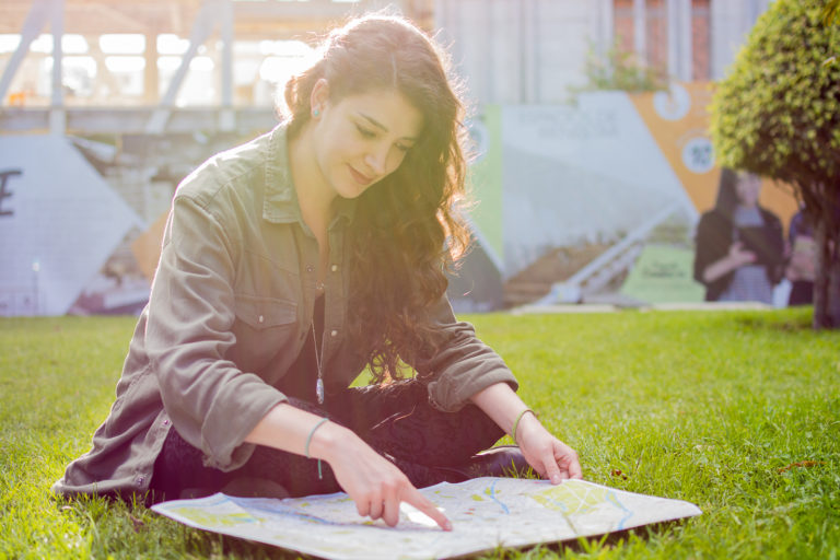 Mujer sentada en el pasto de piernas cruzadas, sonriente, mirando y señalando un mapa mientras cae el sol