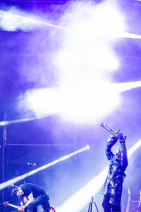 Personaje disfrazado de Infamia mientras guitarrista hace un solo, imagen en tonos fríos
