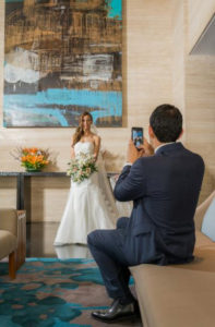 Mujer vestida de novia posando al lado de un cuadro, mientras su esposo le toma una foto.