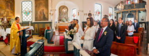 Fotografía panorámica de familia y futuros esposos vestidos de gala de pie, escuchando sermón del sacerdote.