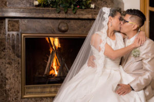 Mujer sentada en las piernas de su pareja, el día de su boda, dandose un beso al lado de la chimenea.