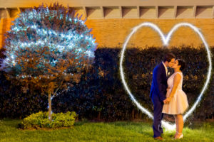 Pareja de esposos besándose con el dibujo de un corazón hecho en luces, al lado de un árbol iluminado