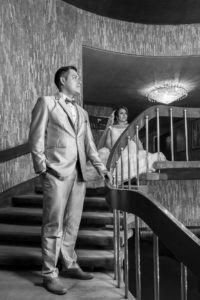 Fotografía en blanco y negro, novio con las manos en el bolsillo y novia sentada en el piso con vestido de novia y velo puesto.